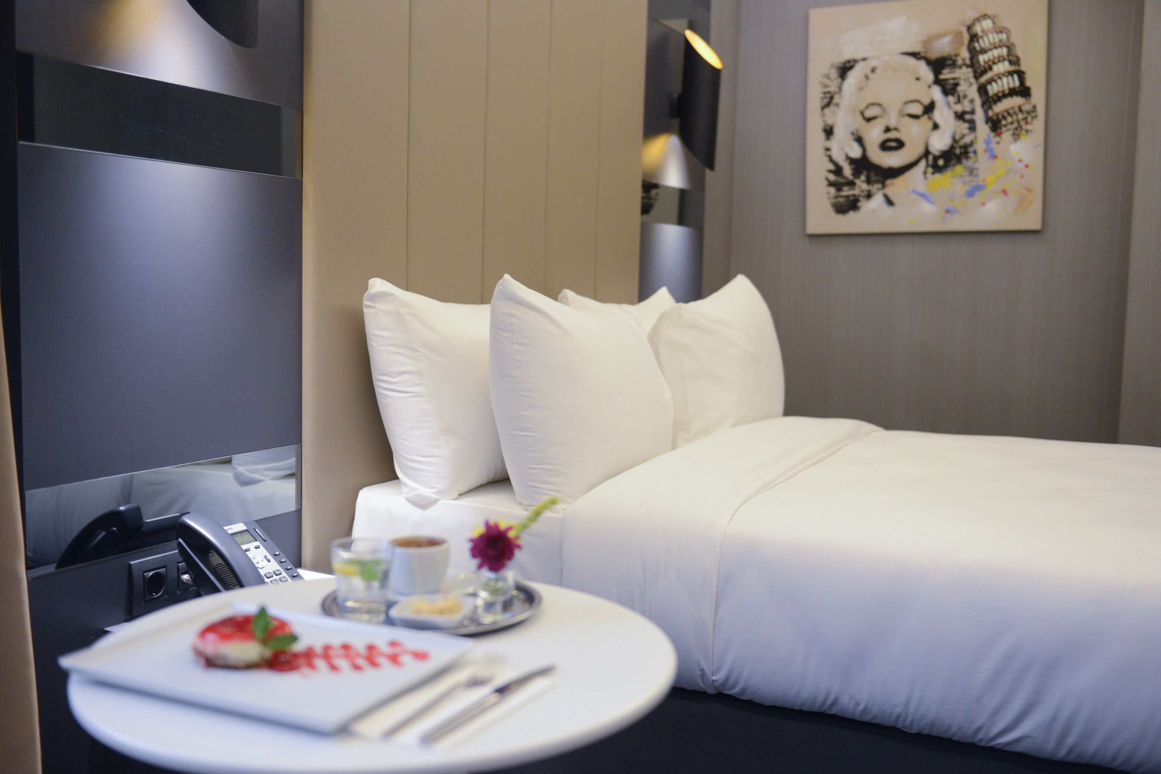 A hotel room with a neatly made bed, a bedside table with a plate of food, a cup, and a vase with flowers.