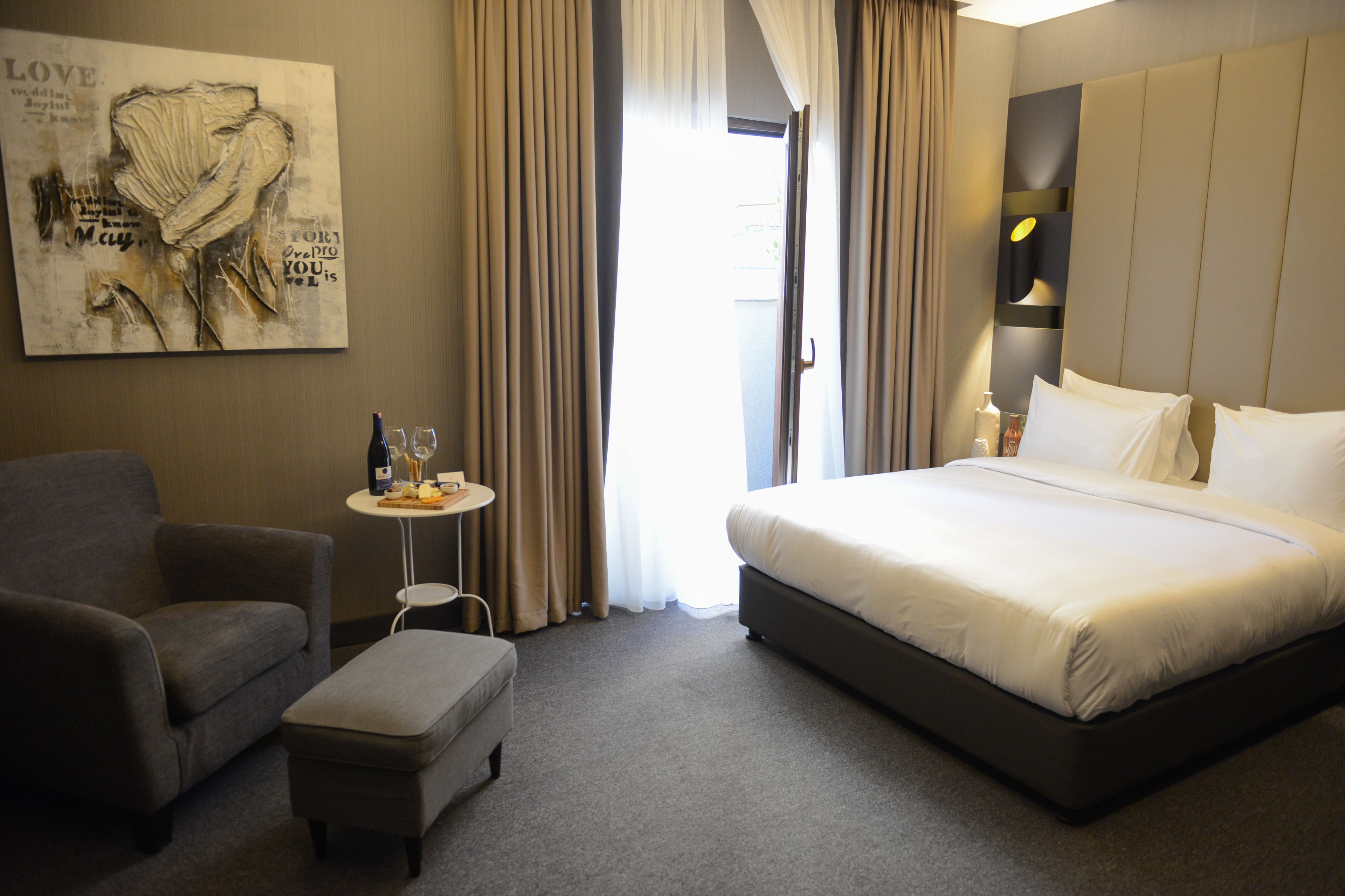 Habitación de hotel con cama, una silla, una mesita con una botella y vasos, y un cuadro en la pared.