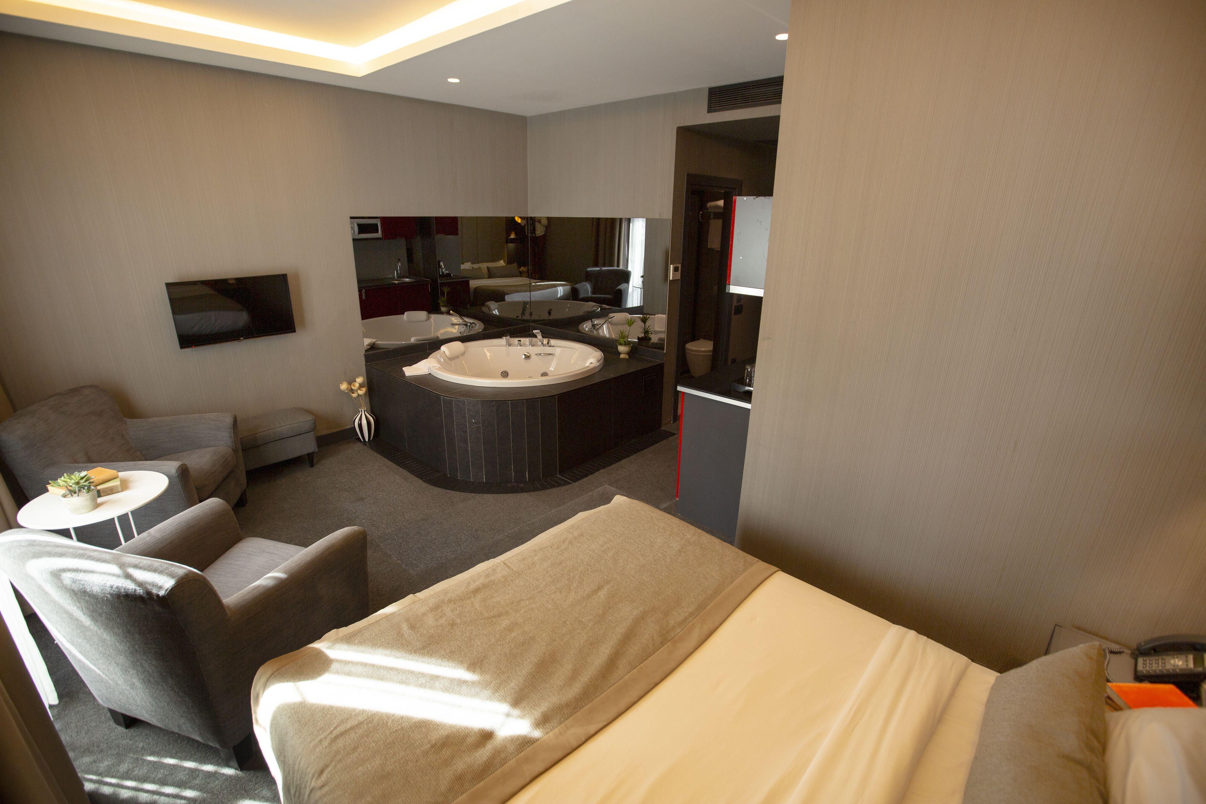 A modern hotel room with a large bed, a sitting area with a sofa and armchairs, a small round table with a plant, and a separate bathroom area with a bathtub and mirror.