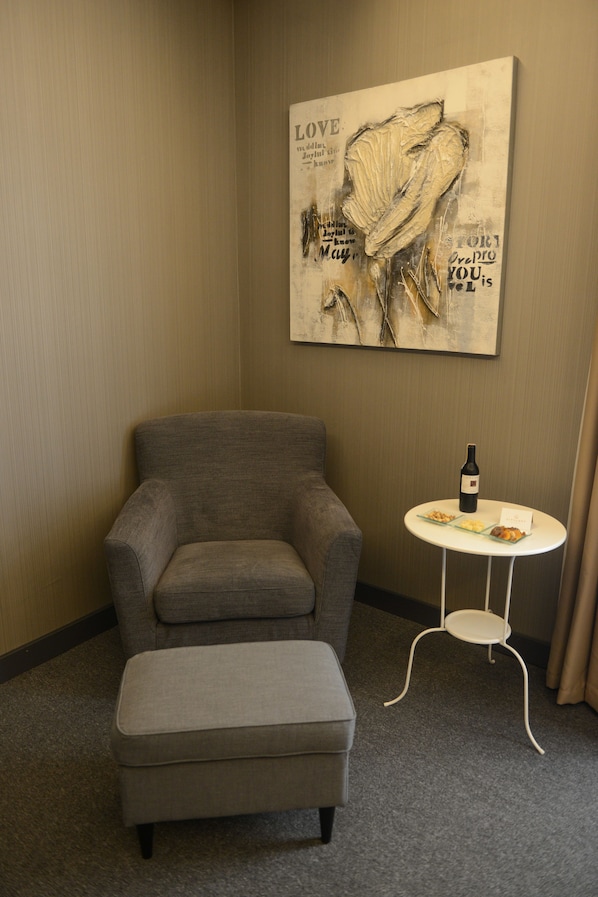 Habitación de hotel con un sillón gris y reposapiés, una mesita redonda con una botella y aperitivos, y una pared con una obra de arte abstracta de gran formato.