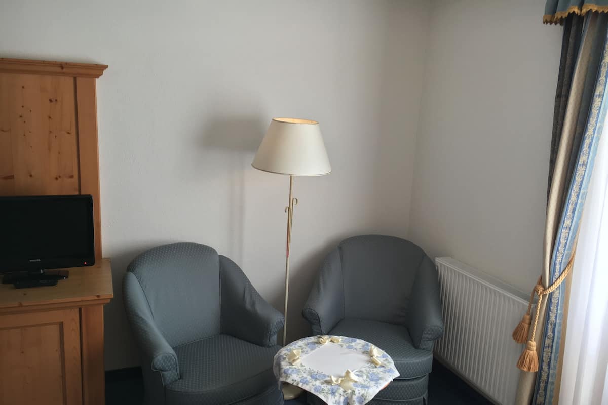 Ein Zimmer mit zwei Sesseln, einem kleinen Tisch mit floraler Tischdecke, einer Stehlampe, einem Fernseher auf einem Holzkasten und einem Fenster mit Vorhängen.