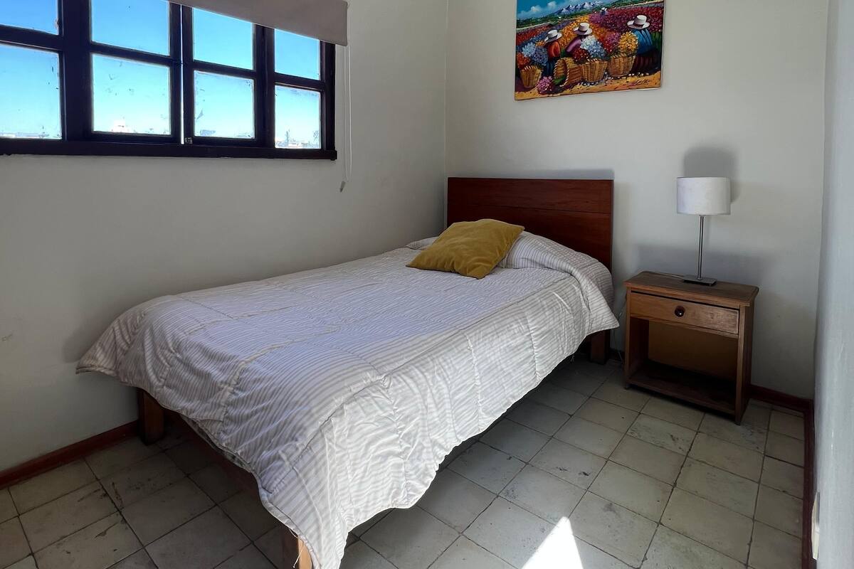 Une chambre à coucher avec un lit, une table de chevet, une lampe, une fenêtre et un tableau au mur.