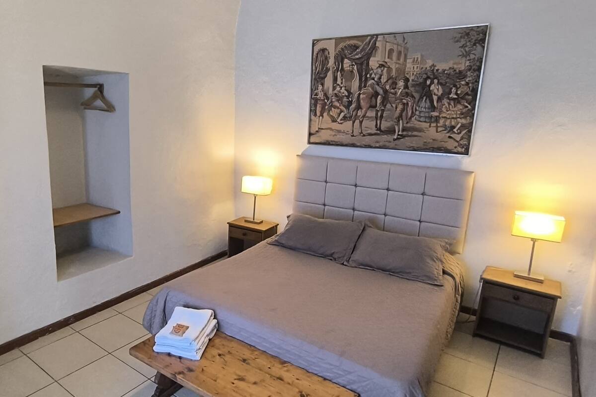 Une chambre à coucher comprenant un lit, des tables de chevet, un porte-manteau mural, une chaise et un tableau encadré accroché au mur.