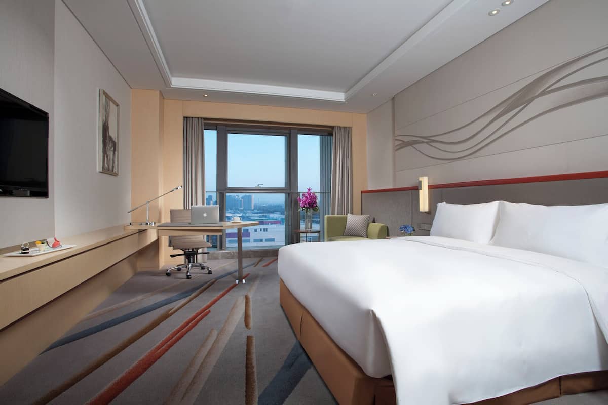 A hotel room with a large bed, a desk with a computer, a TV, and a view of the city through large windows.