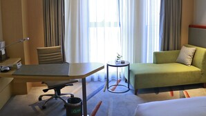 Une chambre d’hôtel avec un lit, un canapé vert, un bureau avec une chaise, une petite table avec une plante et une fenêtre avec des rideaux.
