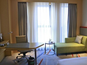 Une chambre d’hôtel avec un lit, un canapé vert, un bureau avec une chaise, une petite table avec une plante et une fenêtre avec des rideaux.
