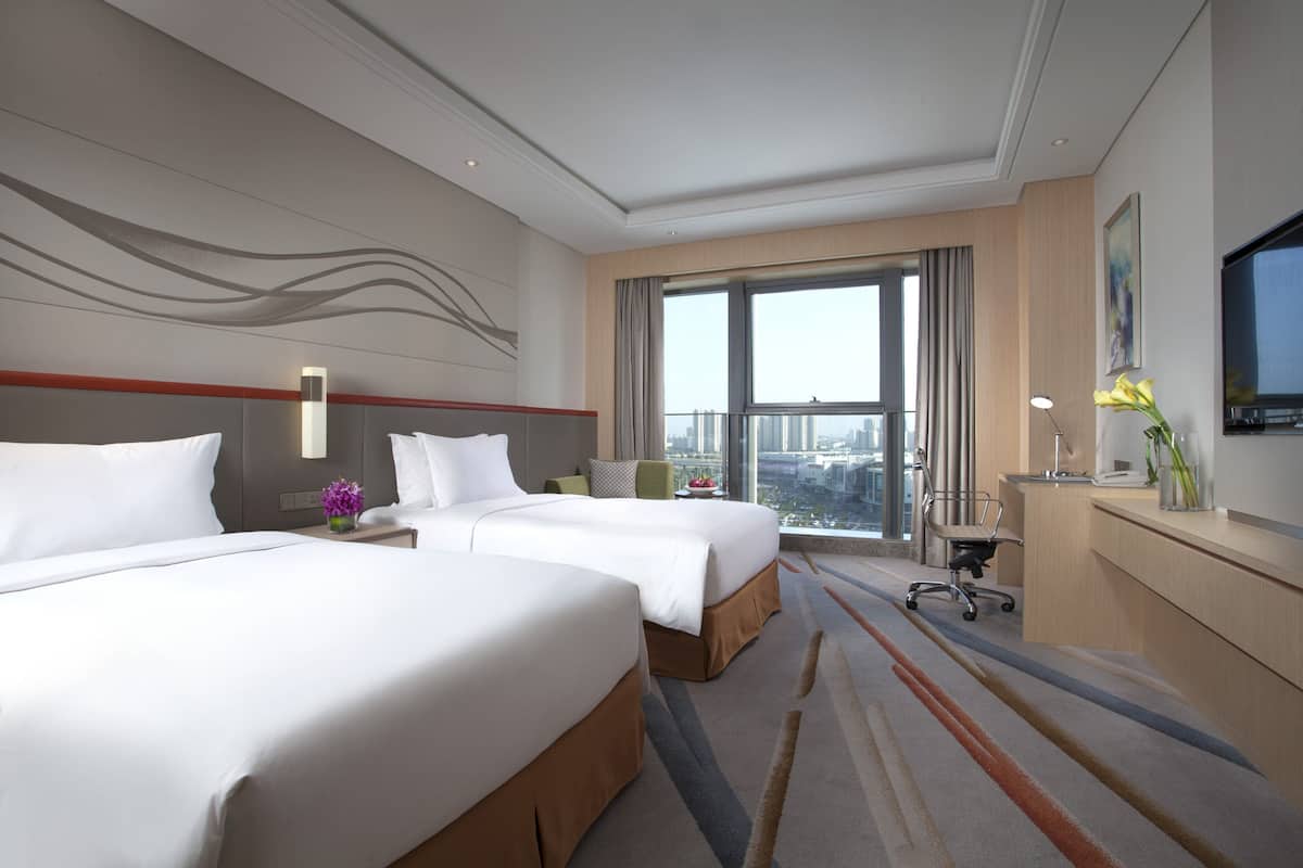 A hotel room with two beds, a desk, a chair, and a large window offering a city view.