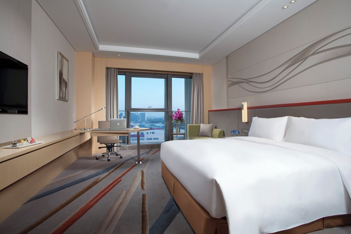 Une chambre d’hôtel dotée d’un grand lit, d’un bureau avec un ordinateur, d’une télévision et offrant une vue sur la ville grâce à de grandes fenêtres.