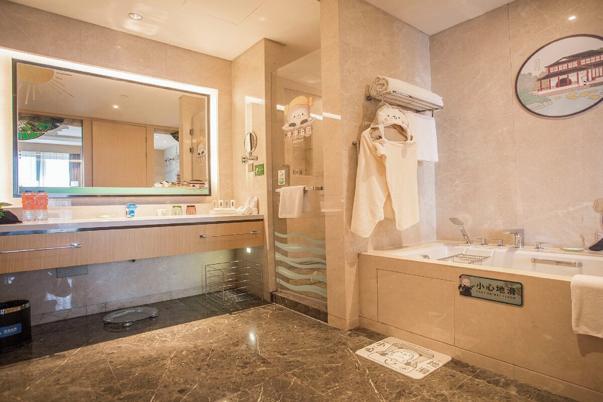 Une salle de bain moderne avec un grand miroir, un lavabo et une baignoire. Des serviettes sont accrochées au mur et un panneau d’avertissement est posé sur le sol.