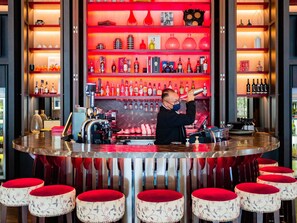 A bar area with a bartender preparing drinks, shelves with bottles, and red cushioned stools.