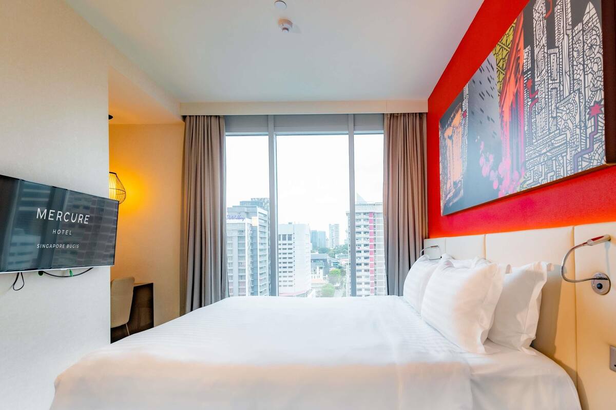 A hotel room with a large bed, a TV mounted on the wall, and a view of the cityscape through the window.