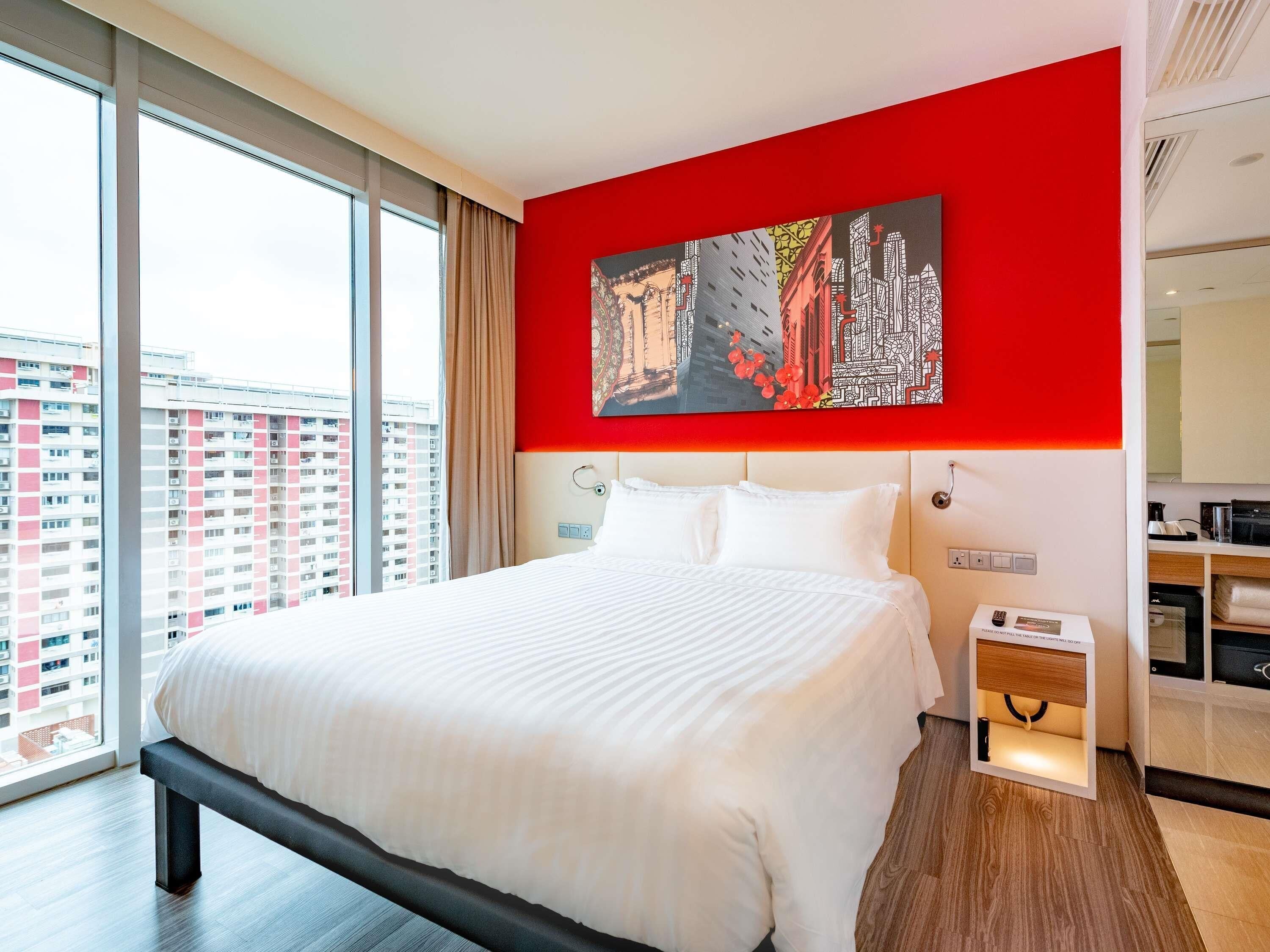 Habitación de hotel con una cama grande, una pared de acento roja, vistas a la ciudad y una mesita de noche con lámpara.