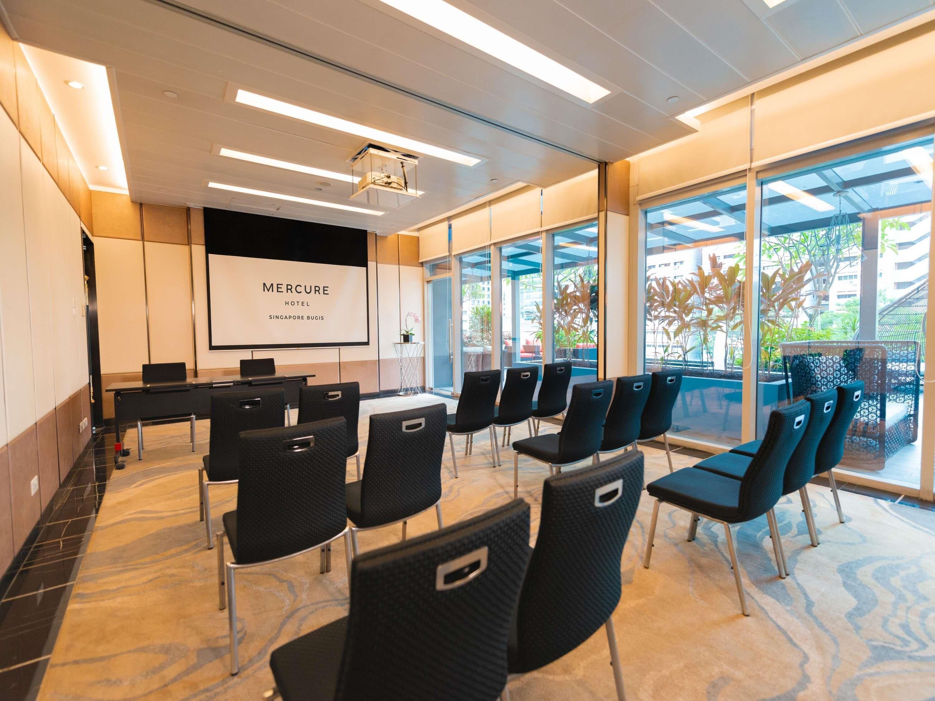 Un salón de conferencias con una pantalla grande mostrando 'Mercure Hotel Singapore Bugis', sillas negras dispuestas alrededor de una mesa y ventanas amplias que ofrecen vistas al exterior.