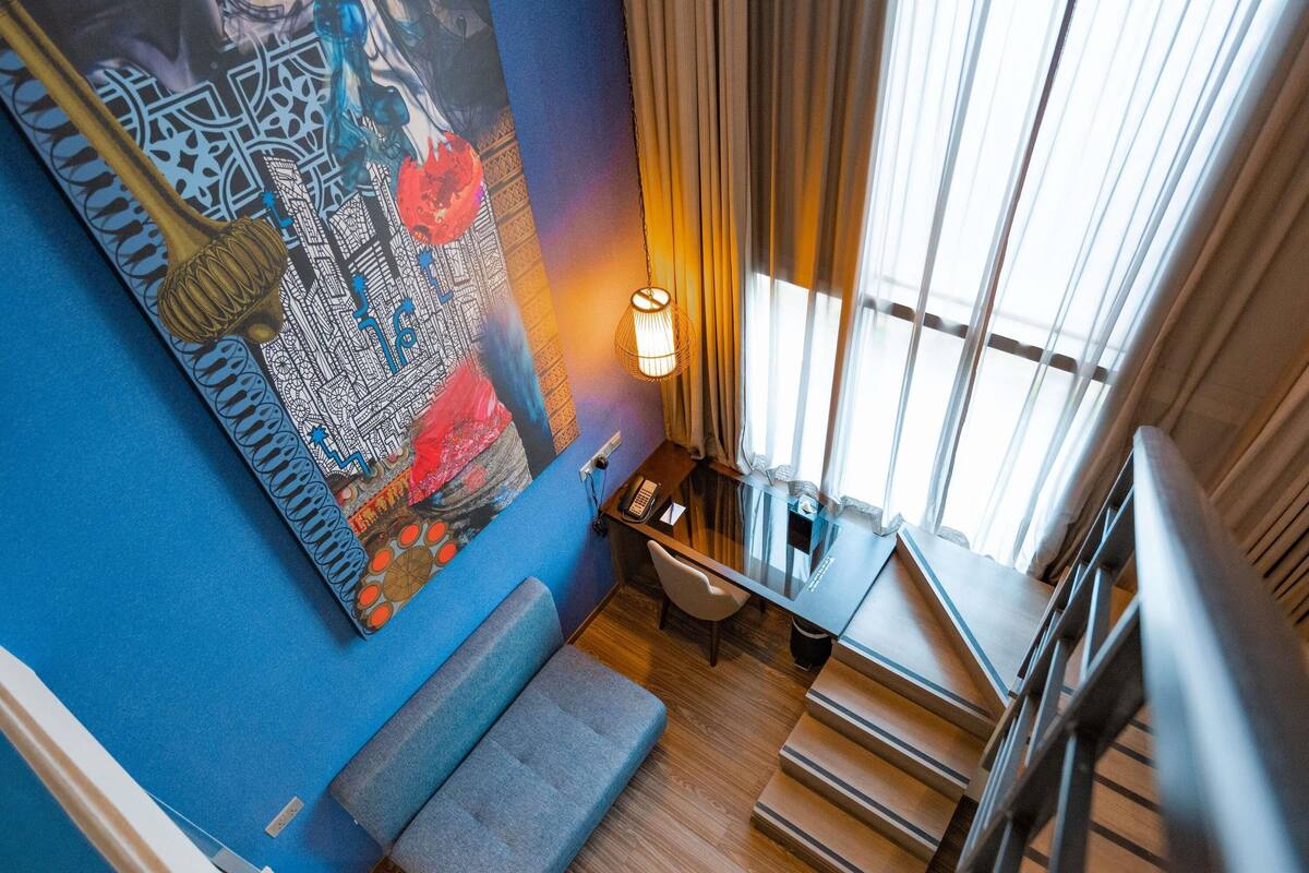 A room with a blue wall, a large abstract painting, a desk with a lamp, a staircase, and a window with curtains.