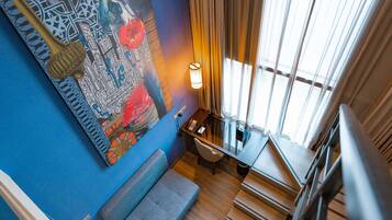 Ein Zimmer mit blauer Wand, einem großen abstrakten Gemälde, einem Schreibtisch mit Lampe, einer Treppe und einem Fenster mit Vorhängen.