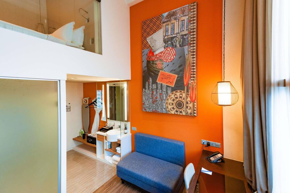 A modern hotel room with a blue sofa, a large wall art piece, a hanging lamp, and a small desk with a telephone.