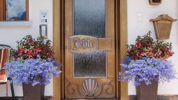 En bygningseingang med et skilt hvor der står "Jehle" og to store blomsterkrukker med røde og blå blomster.