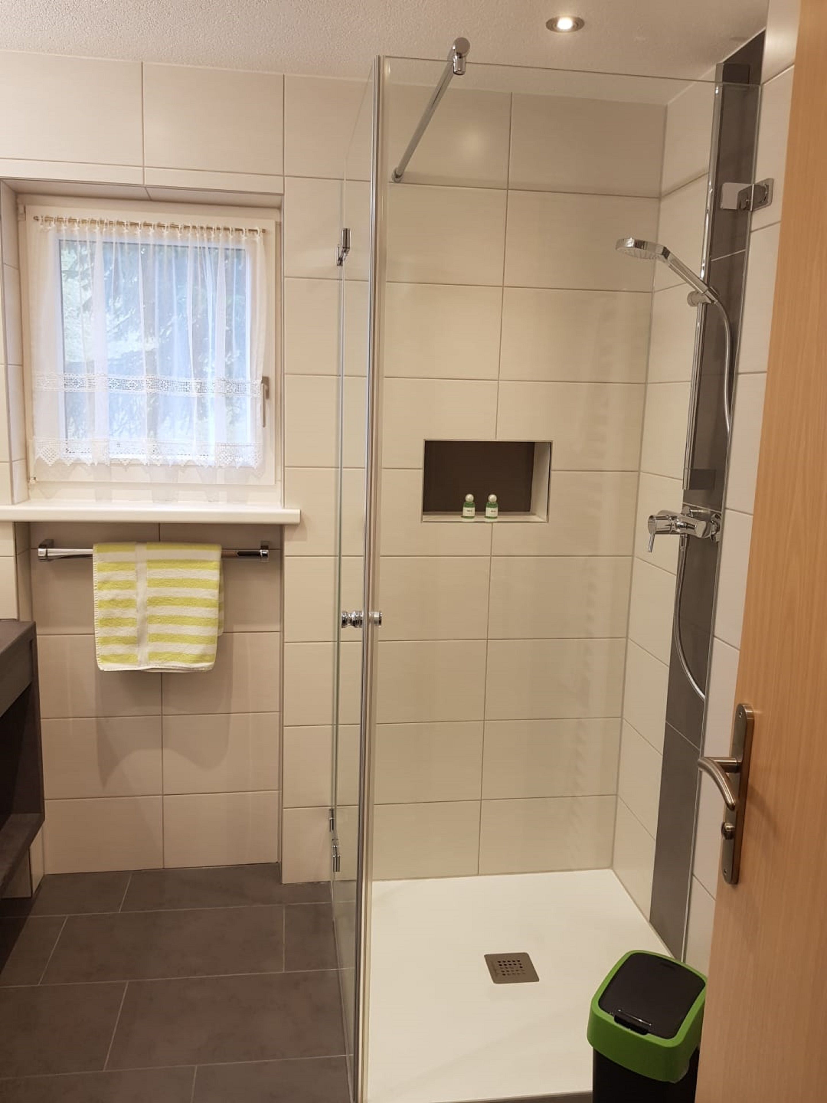 A modern bathroom with a glass shower enclosure, a window with a lace curtain, and a towel rack with a striped towel.
