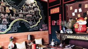A room with a blackboard wall featuring Chinese calligraphy and drawings of landmarks. There is a traditional tea set on a table, a small altar with incense, and a decorative red figure. - Tomato Red (Jinsha)