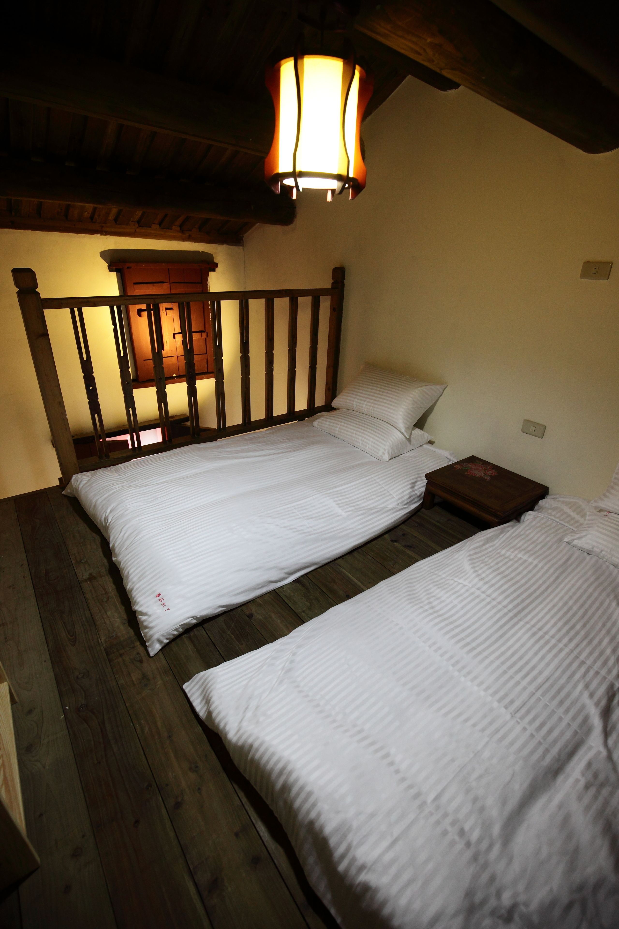 A room with two single beds, wooden flooring, a wooden headboard, a nightstand, and a wall-mounted lamp.