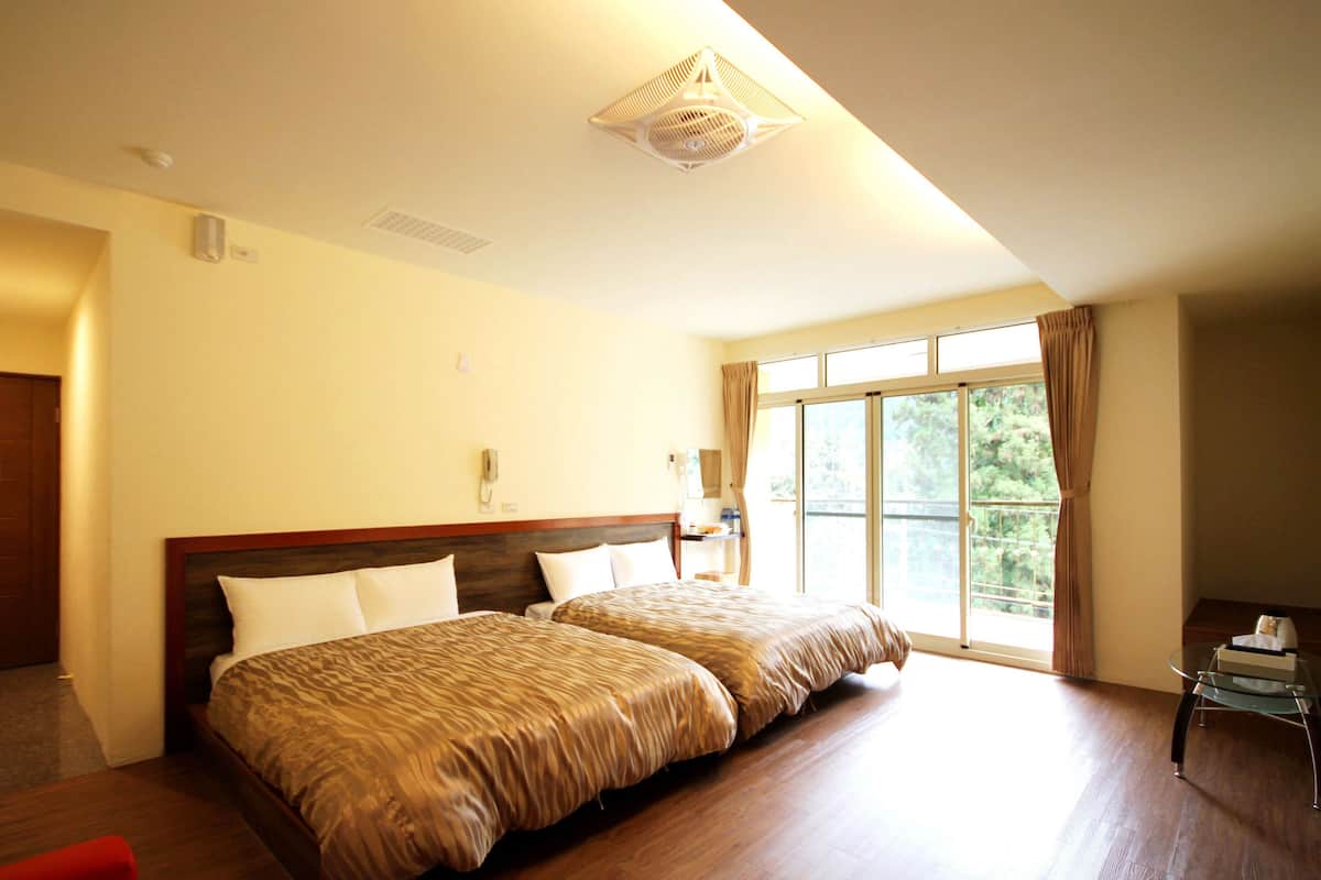 A hotel room with two beds, a small table, a chair, a window with curtains, and a ceiling light fixture.