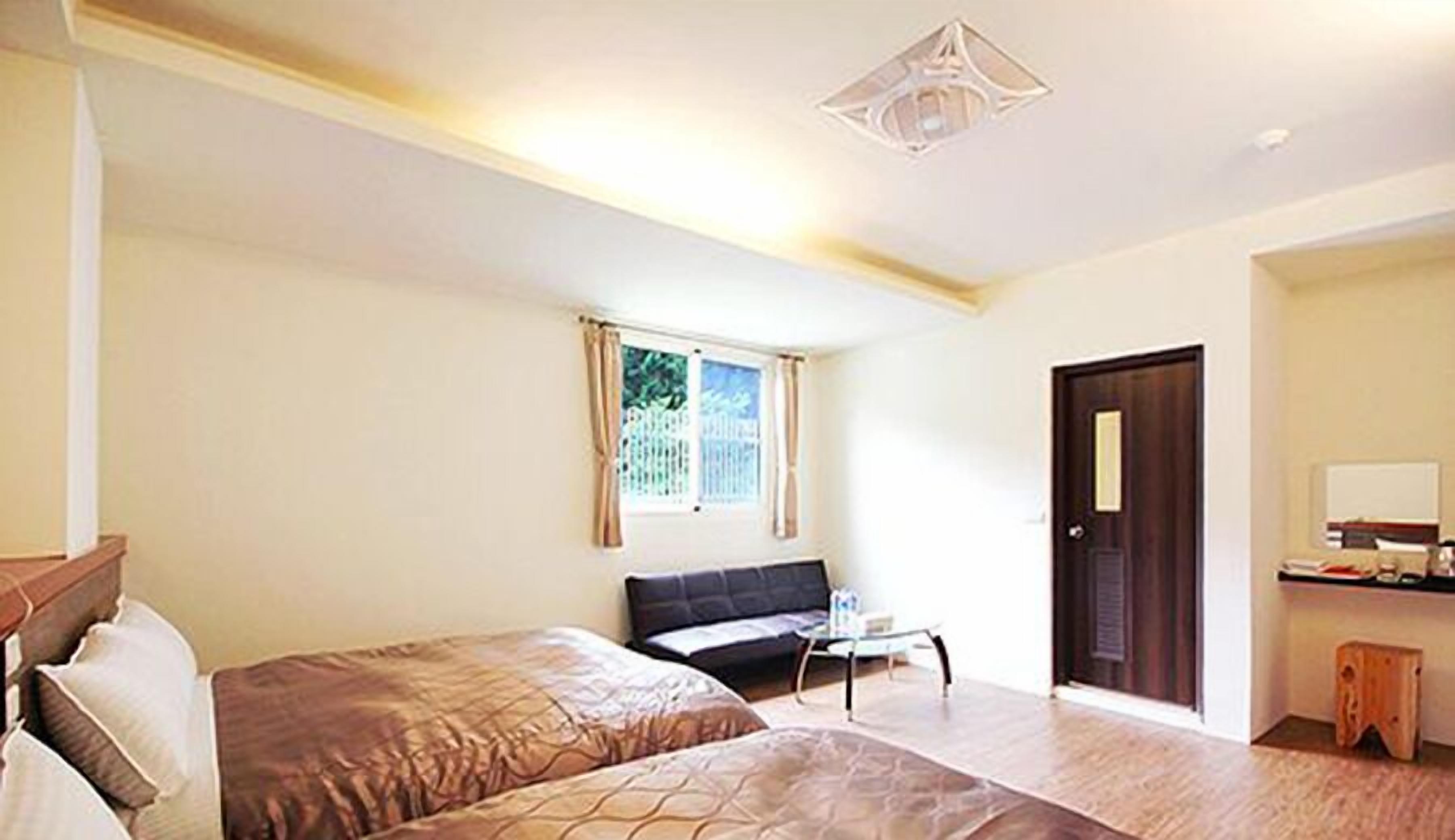 A room with two beds, a small table, a window with curtains, a door, and a ceiling light fixture.