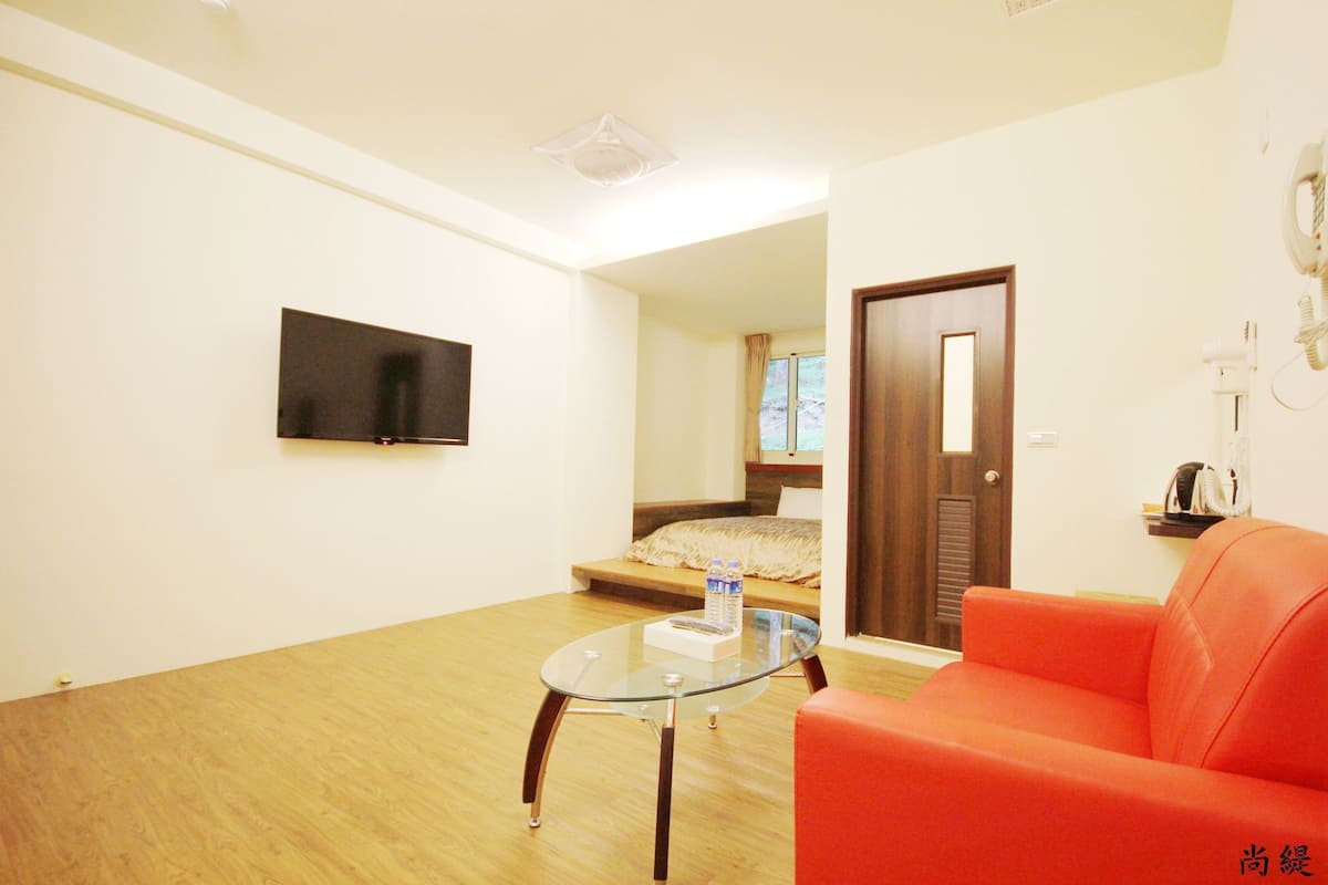 A modern living room with a glass coffee table, a red sofa, a flat-screen TV mounted on the wall, and a wooden floor.