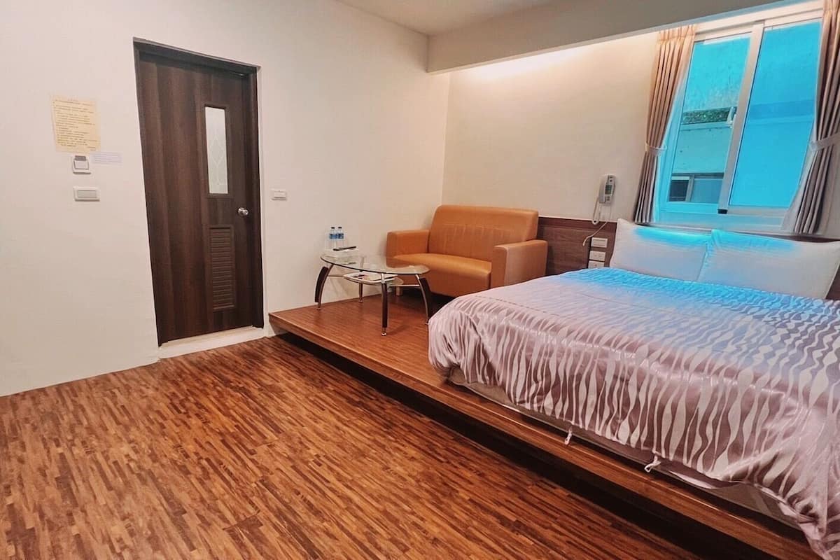 A hotel room with a bed, a chair, a small table, a door, and a window with curtains.