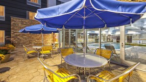 Eine Terrasse im Freien mit gelben Metallstühlen und einem runden Tisch, beschattet durch einen blauen Sonnenschirm.