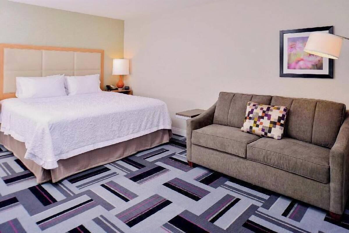 A hotel room with a bed, a sofa, a nightstand, a lamp, and a framed picture on the wall.