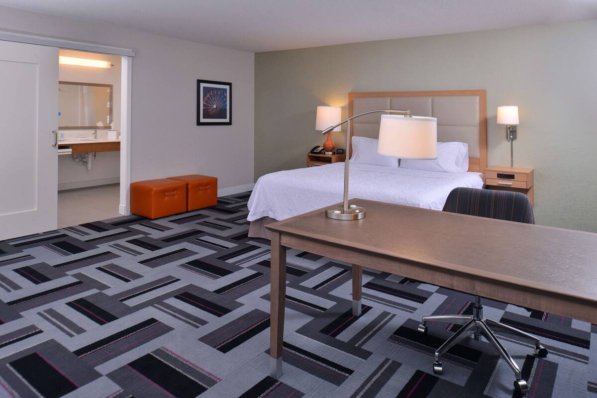 A hotel room with a bed, bedside tables, a desk, an orange ottoman, and a patterned carpet.