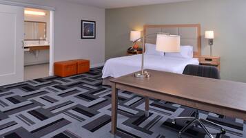 A hotel room with a bed, bedside tables, a desk, an orange ottoman, and a patterned carpet.