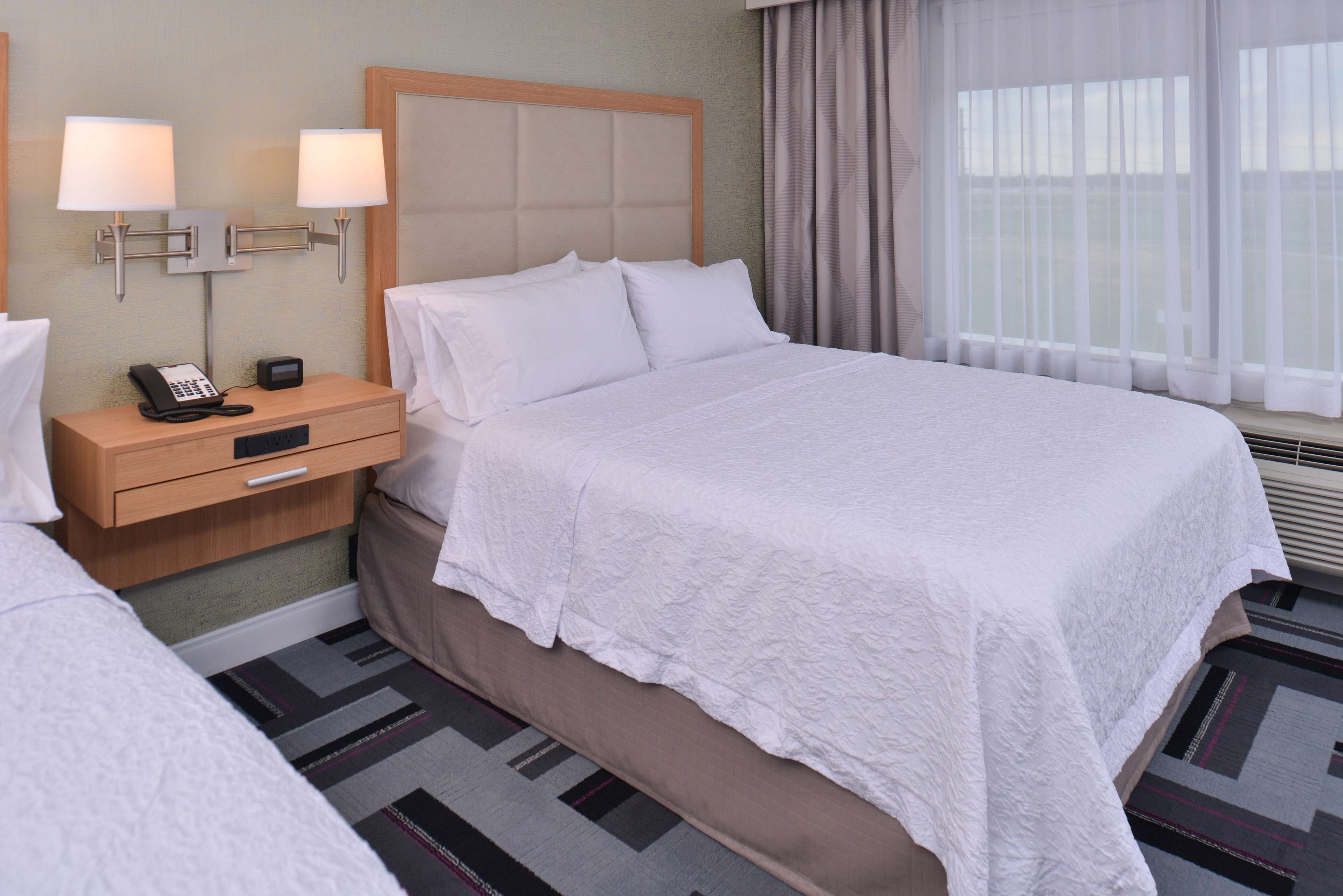 A hotel room with a large bed, a nightstand with a phone and a lamp, and a window with curtains.