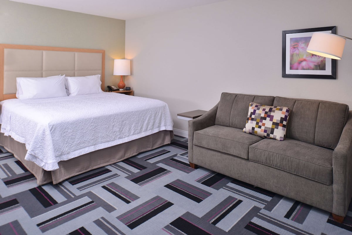 A hotel room with a bed, a grey sofa, a patterned rug, a nightstand with a lamp, and a framed picture on the wall.