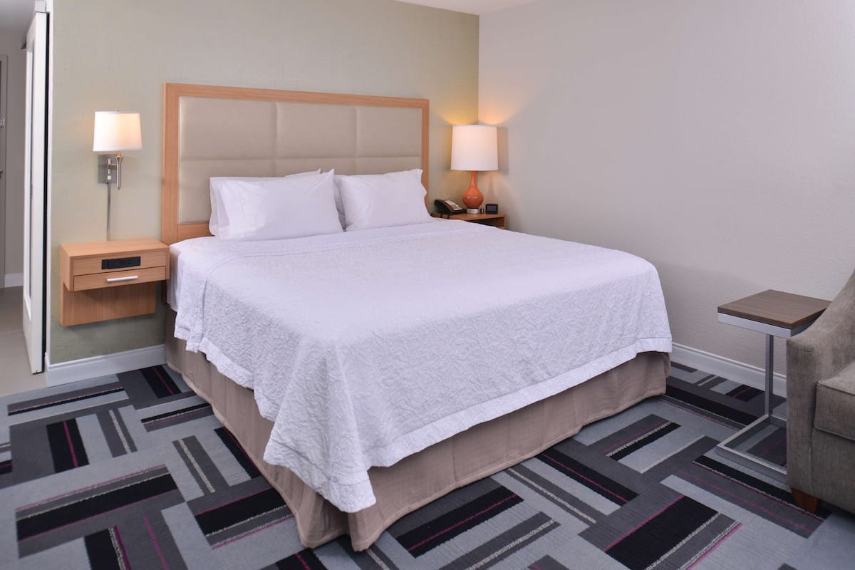 A neatly made bed with a white bedspread and pillows, a wooden nightstand with a lamp, and a small table with a lamp.