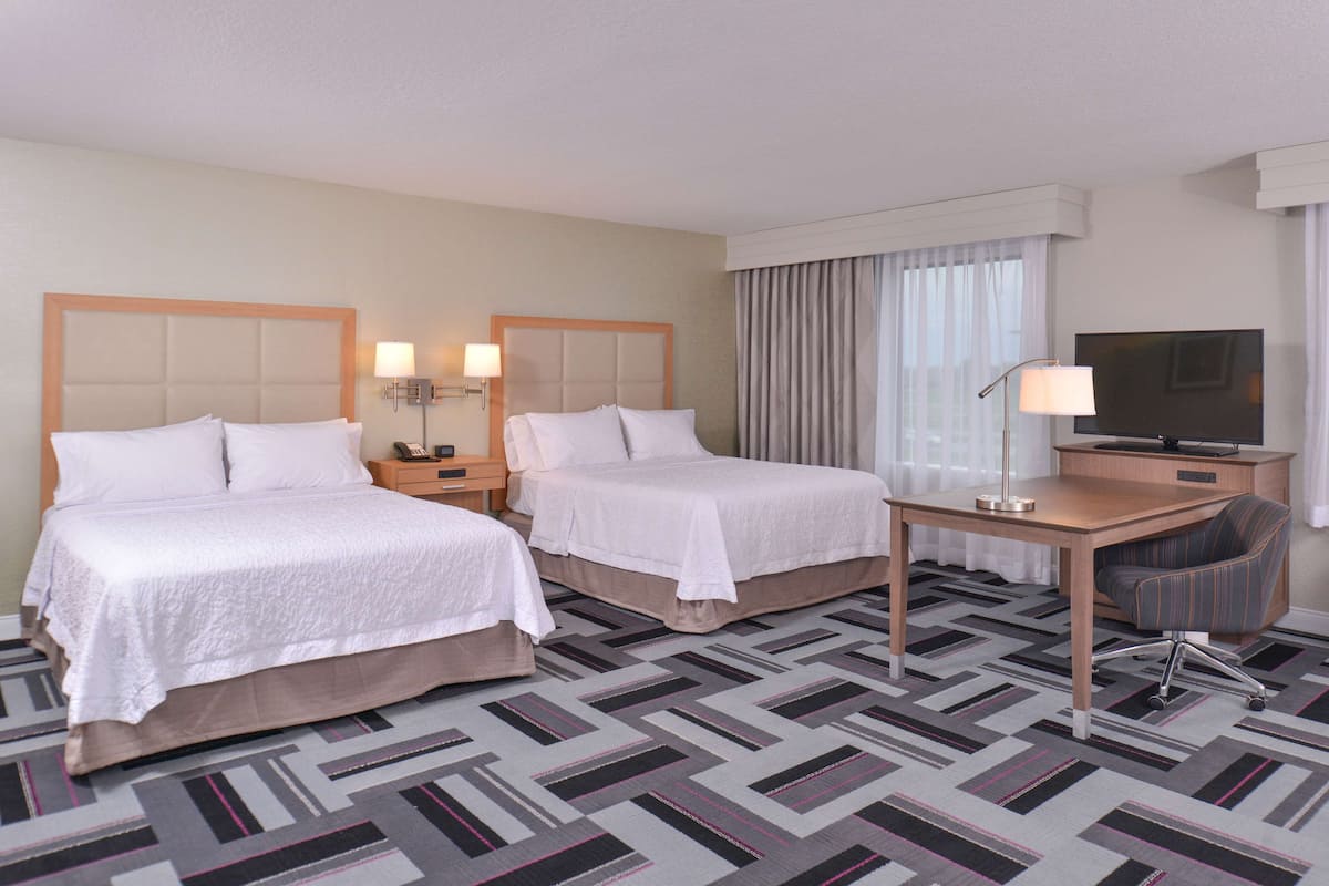 A hotel room with two beds, a desk, a chair, a TV, and a lamp.