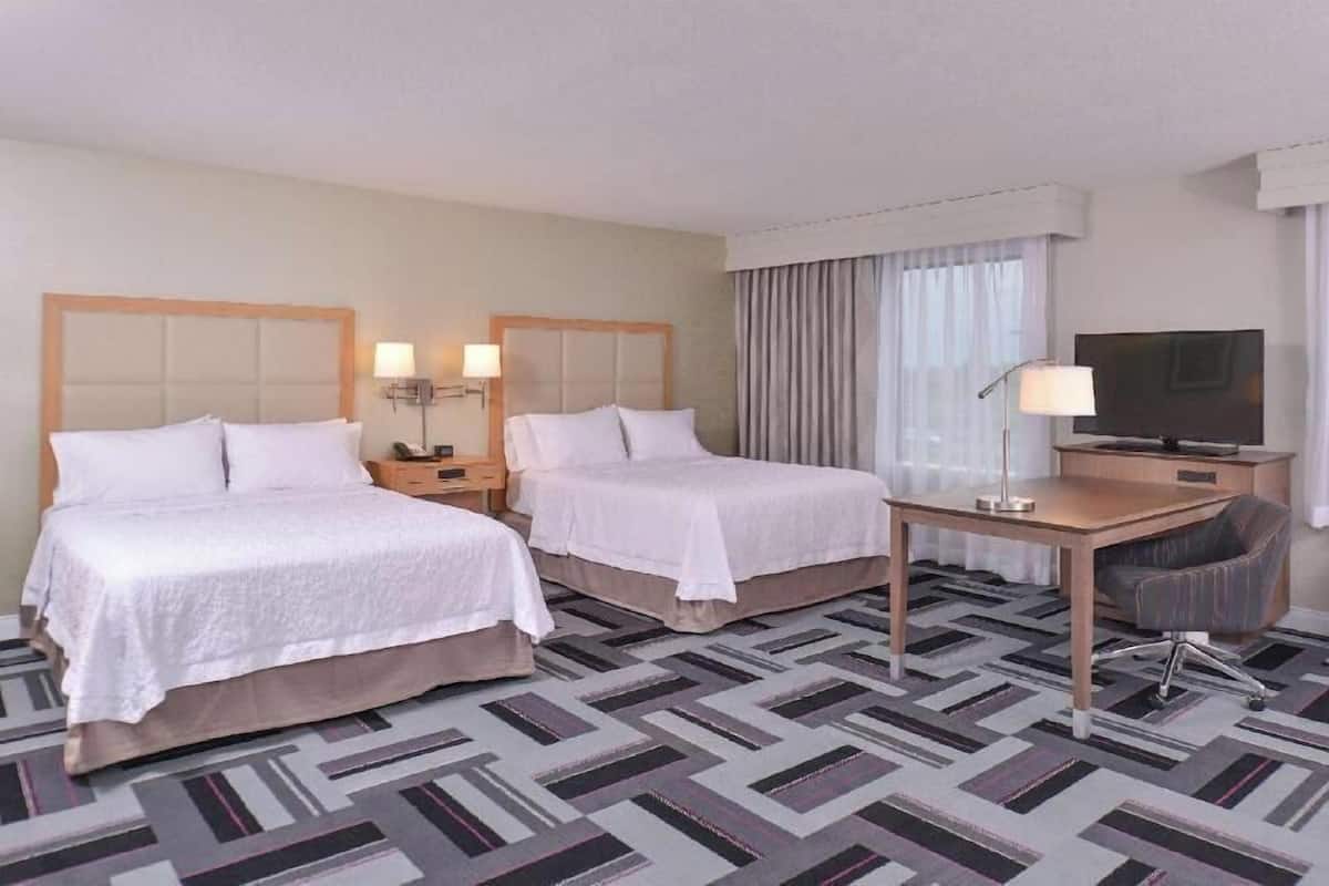 A hotel room with two beds, a desk, a chair, a television, and a lamp.