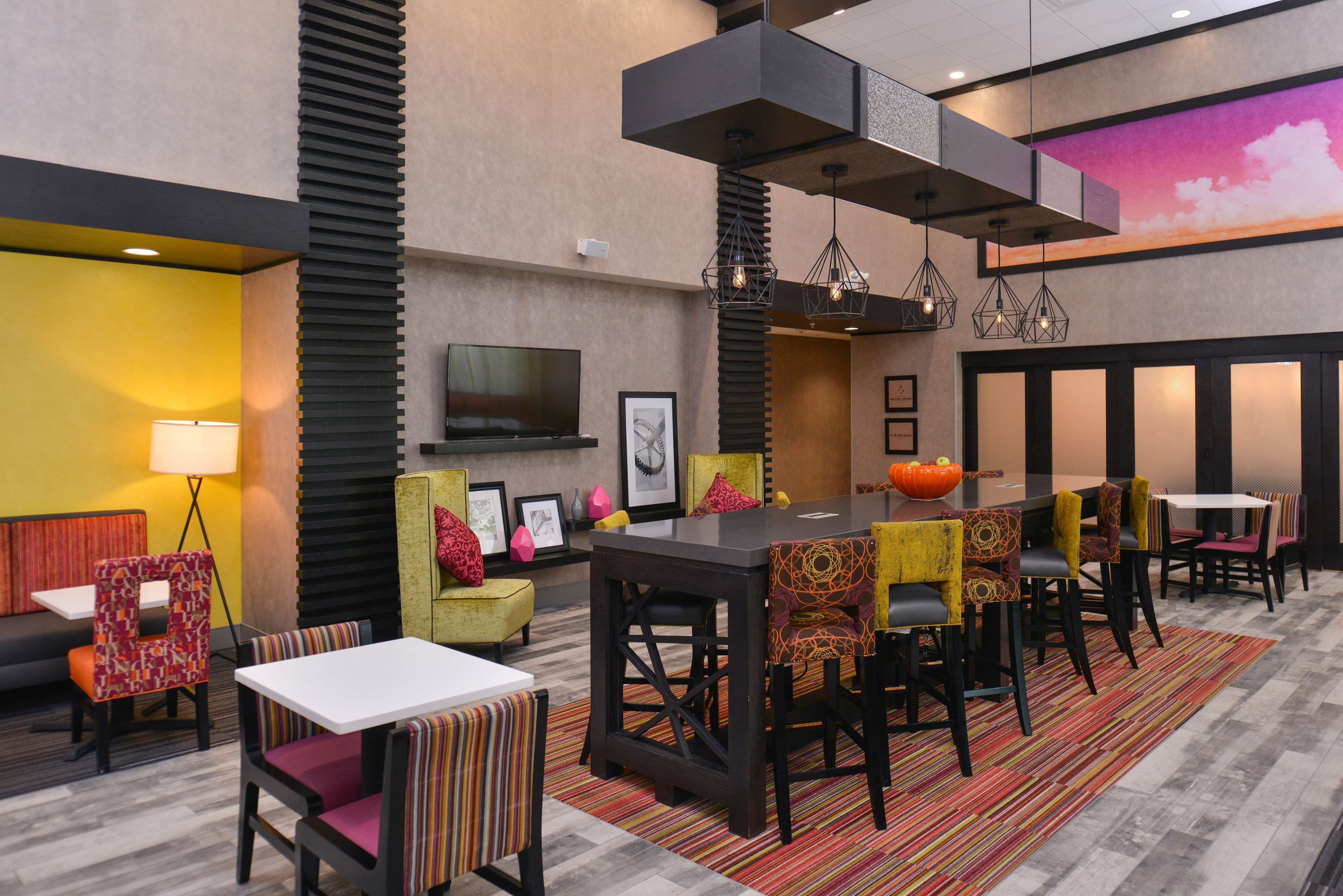 A modern hotel lobby with a large dining table, comfortable seating, a flat-screen TV, and a colorful rug.
