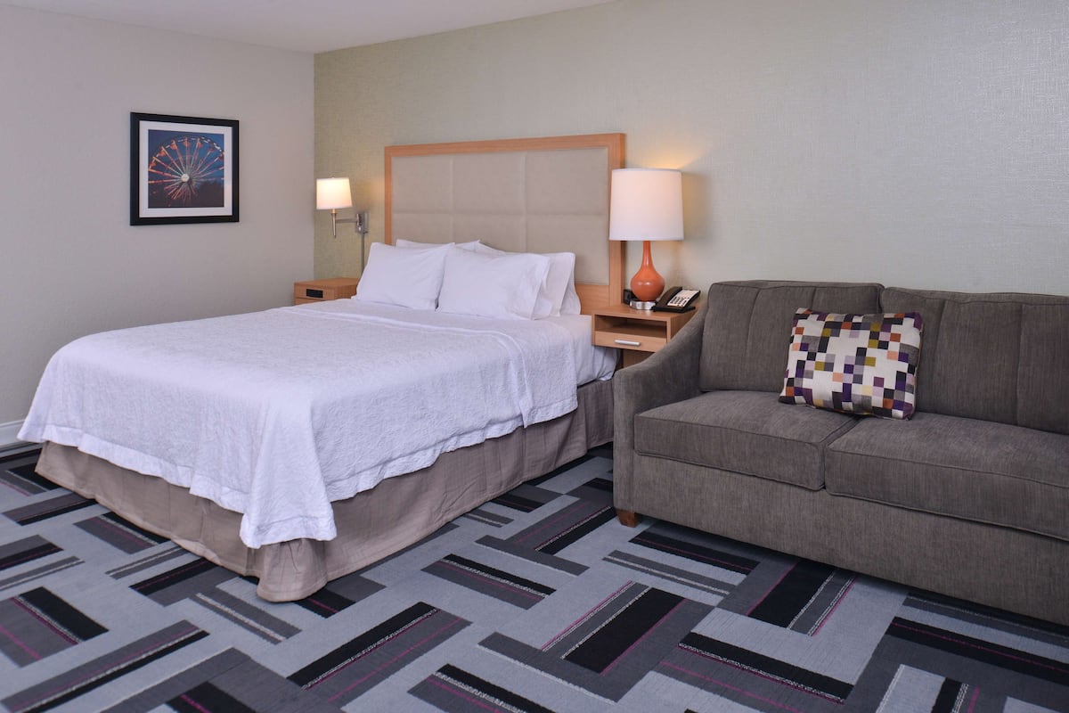 A hotel room with a bed, a sofa, a nightstand with a lamp, and a framed picture on the wall.
