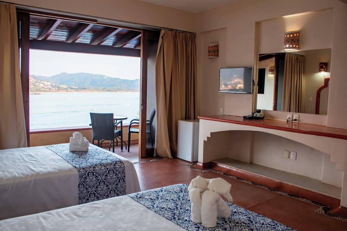 A hotel room with two beds, a desk, a chair, a TV, and a view of the sea.