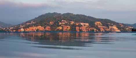 A coastal cityscape with buildings reflecting on calm water, a prominent hill with structures, and a clear sky.
