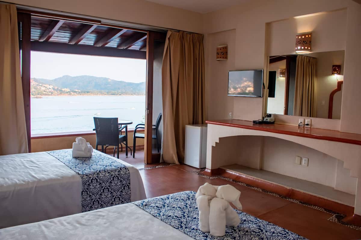 A hotel room with two beds, a desk, a chair, a TV, and a view of the sea.