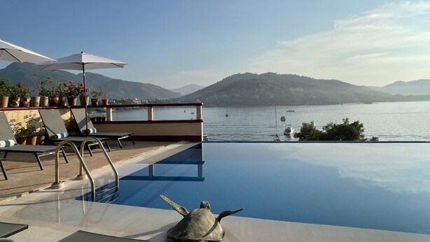 Una piscina infinita con vista a un lago y montañas, sillas de descanso y plantas en macetas.