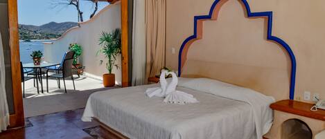 Quarto com cama, cabeceira de madeira, uma mesa pequena com cadeiras e vista para o mar.