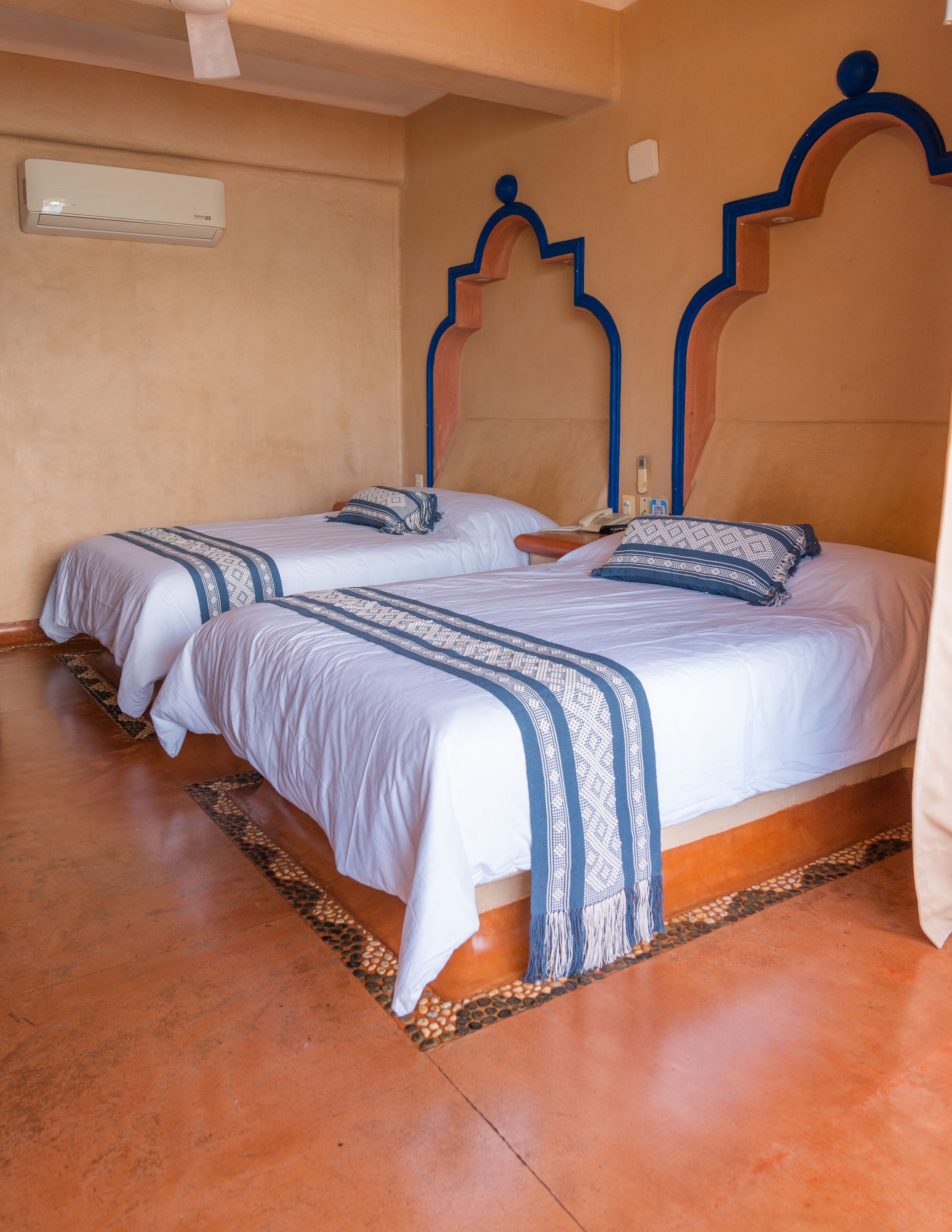 Ein Zimmer mit zwei Betten, jeweils mit gemustertem Bettbezug und Kissen, einem Deckenventilator und einer Wand-Klimaanlage.