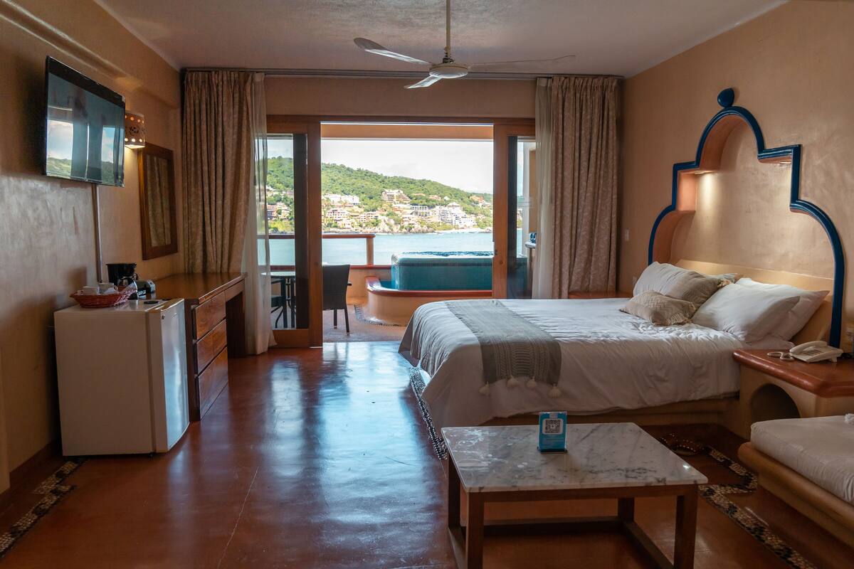 A hotel room with a bed, a desk, a chair, a small table, a balcony with a view of the sea, and a ceiling fan.