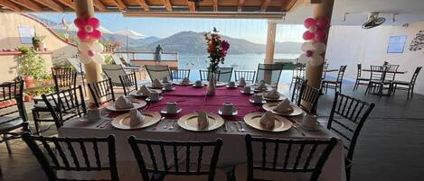 Un área de comedor al aire libre con vistas al mar y a las montañas, que cuenta con techo de madera, mesas redondas con servilletas blancas y sillas negras.