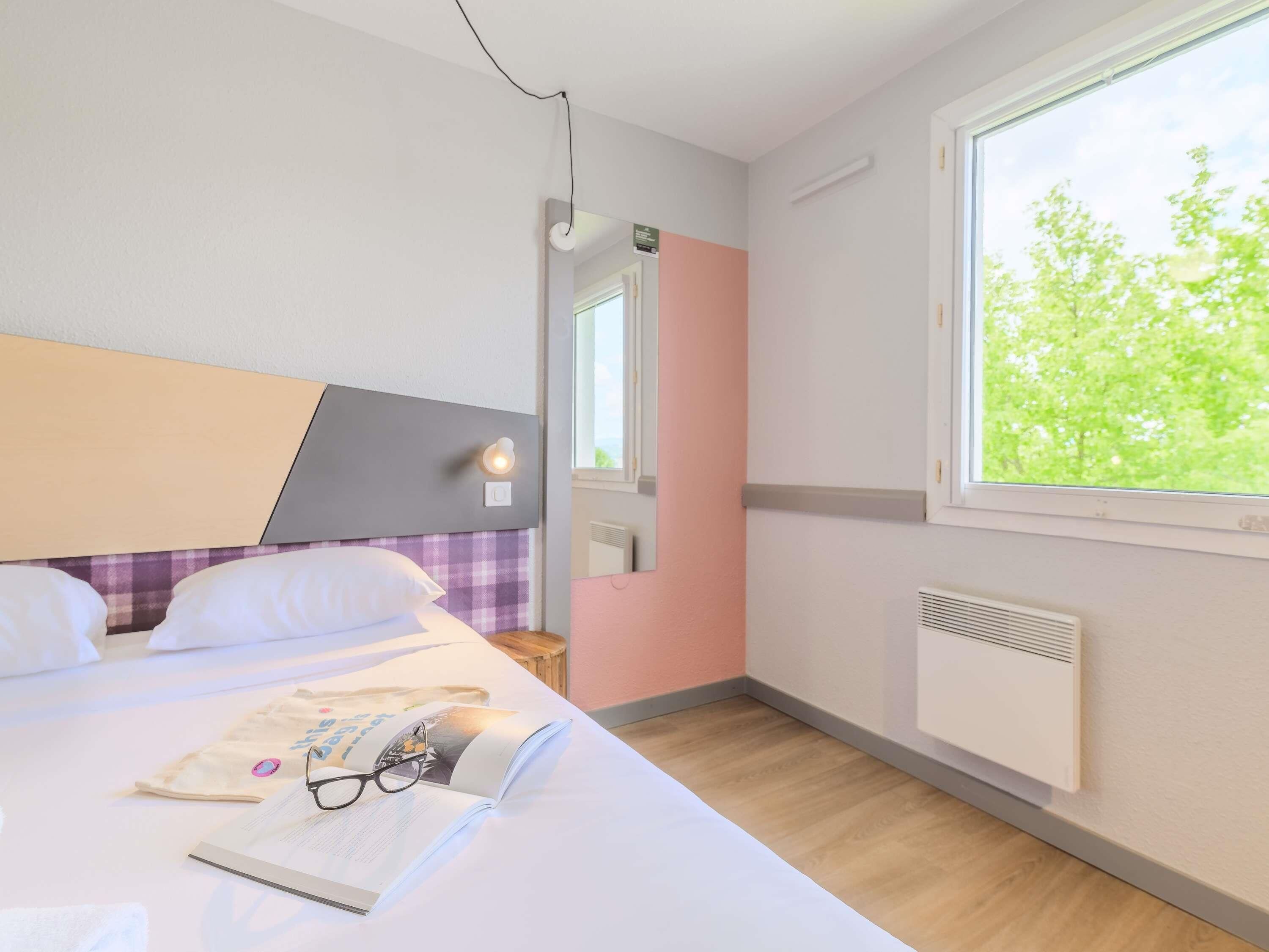 A bedroom with a bed, a window with a view of trees, a bedside table with a book and glasses, and a wall-mounted heater.