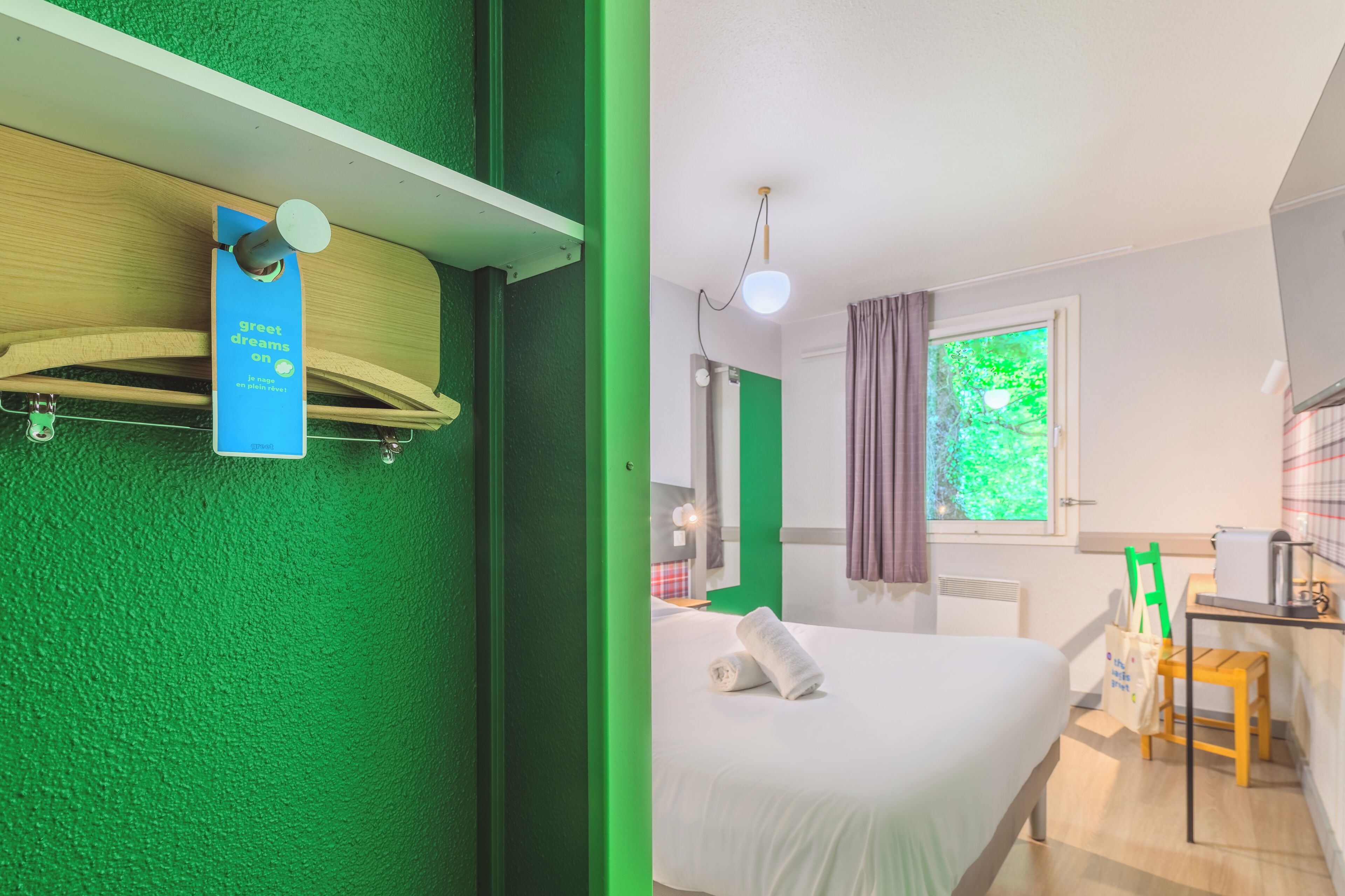 A hotel room with a green wall, a bed with a white comforter, a small table with a chair, a window with a view of greenery, and a ceiling light fixture.