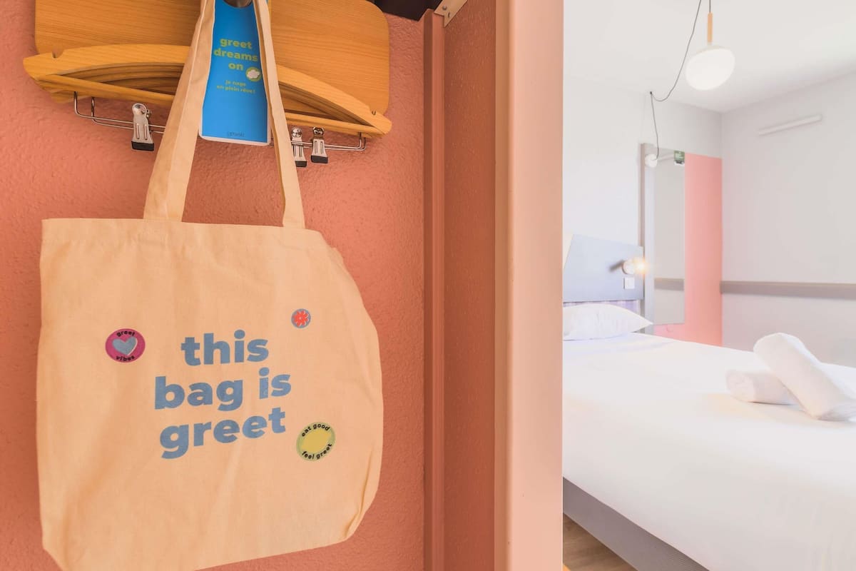 A hotel room with a bed, a nightstand, a lamp, and a wall-mounted shelf with a book and a tote bag.